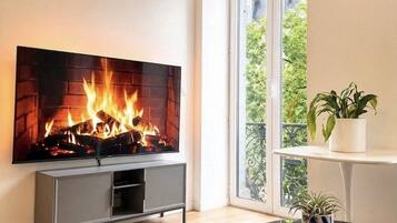 Una sala de estar con un televisor de pantalla plana montado en la pared, una chimenea con leña, un mueble de entretenimiento gris, una mesa de centro redonda de vidrio y una planta en maceta sobre un estante blanco.