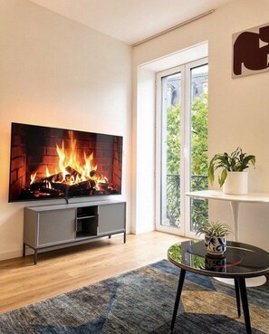 Ett vardagsrum med en platt-TV som är monterad på väggen, en öppen spis med ved, en grå TV-bänk, ett runt soffbord i glas och en krukväxt på en vit hylla.
