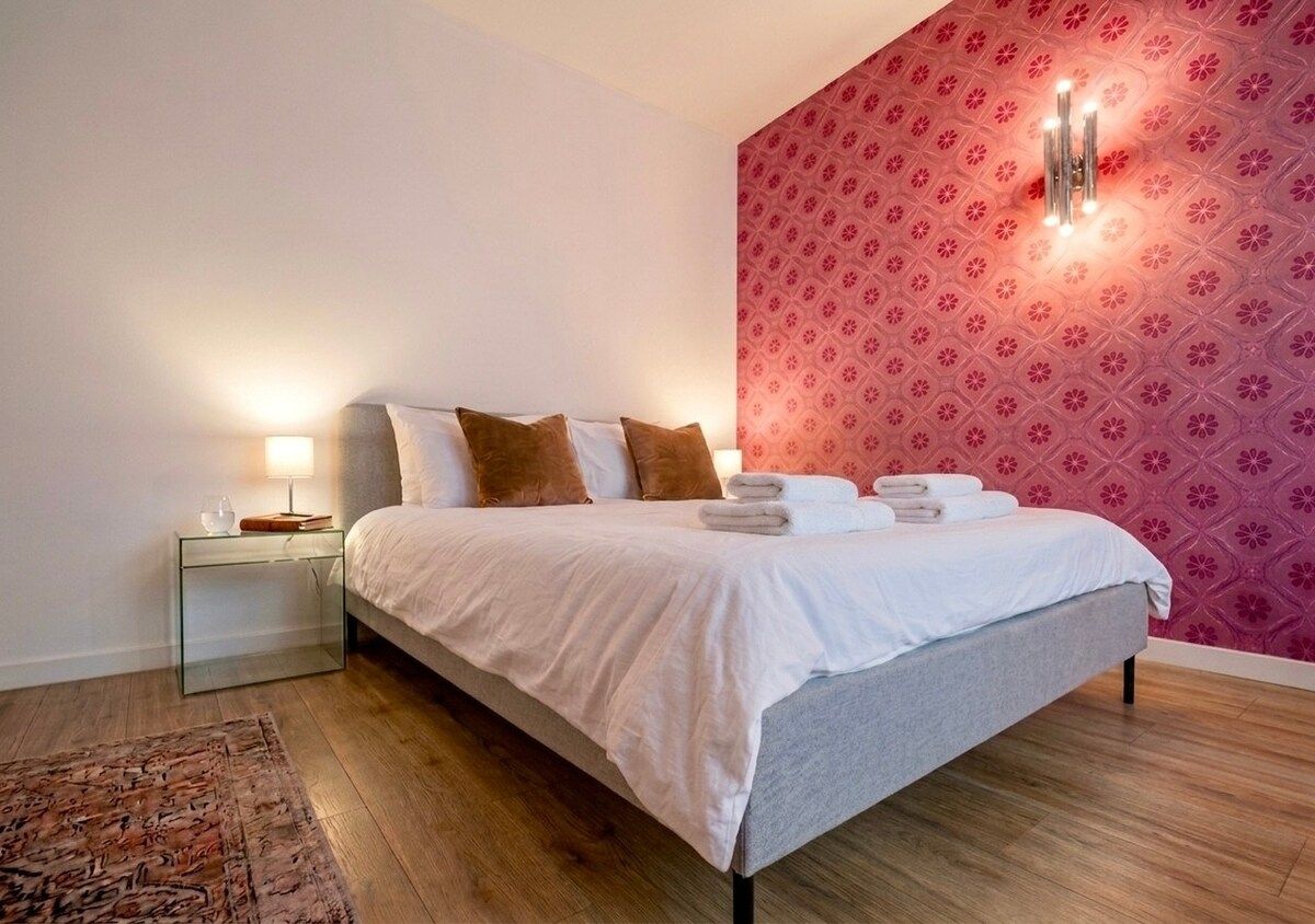 Una cama bien tendida con ropa de cama blanca y gris, una mesita de noche de cristal con una lámpara, y un papel tapiz estampado al fondo.