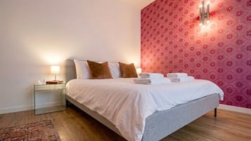 Una cama bien tendida con ropa de cama blanca y gris, una mesita de noche de cristal con una lámpara, y un papel tapiz estampado al fondo.
