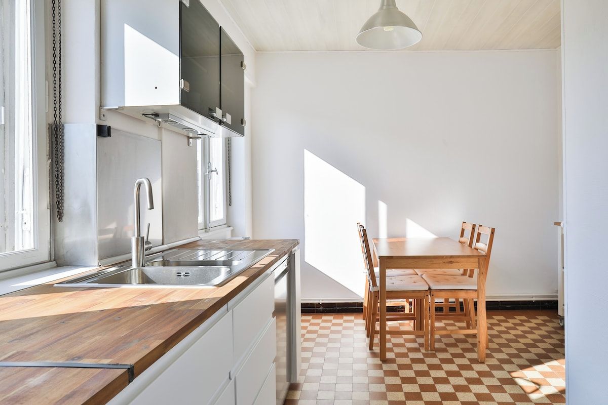 Una cocina con encimera de madera, pileta de acero inoxidable y comedor con piso a cuadros, mesa y sillas.