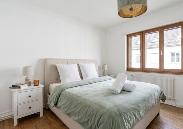 Un dormitorio con cama, mesita de noche, lámpara y ventana con vistas a edificios.