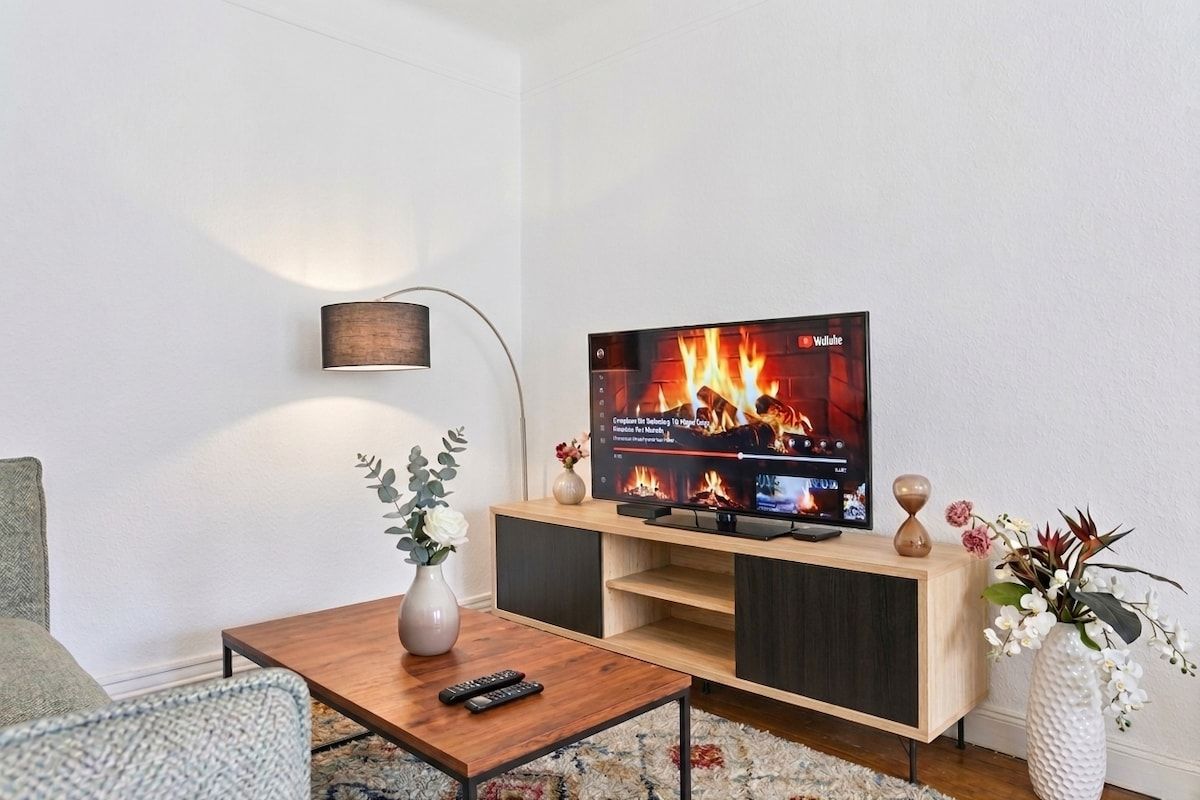 Una sala de estar moderna con un televisor sobre un mueble de madera, un florero con flores y un control remoto en una mesa de centro.