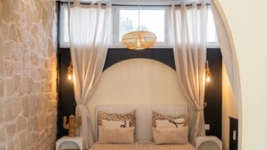 Quarto com parede de pedra, cama com roupa de cama estampada, luminária pendente e janela com cortinas.