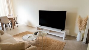 Sala de estar com televisão de tela plana, sofá, mesa de centro e área de jantar com cadeiras.