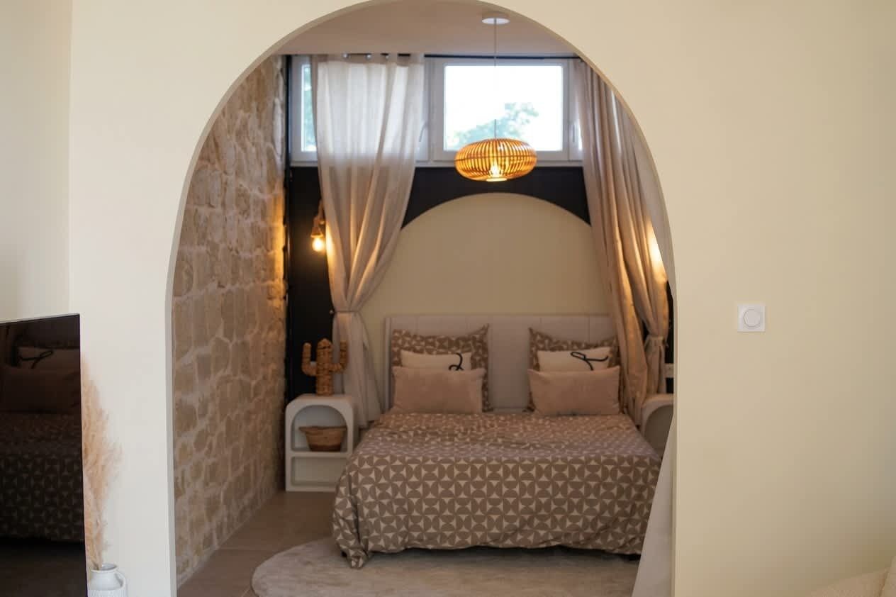 Quarto com arco de pedra, cama com roupa de cama estampada, luminária pendente e janela com cortinas.
