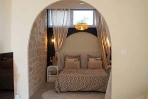 A bedroom with a stone archway, a bed with patterned bedding, a hanging light fixture, and a window with curtains.