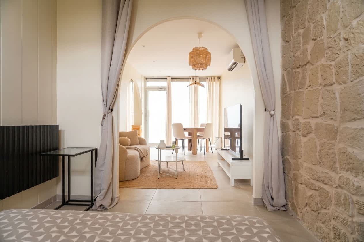 Un salon moderne avec un canapé, une table basse et un coin repas avec des chaises. On y trouve un mur en pierre et une grande fenêtre.