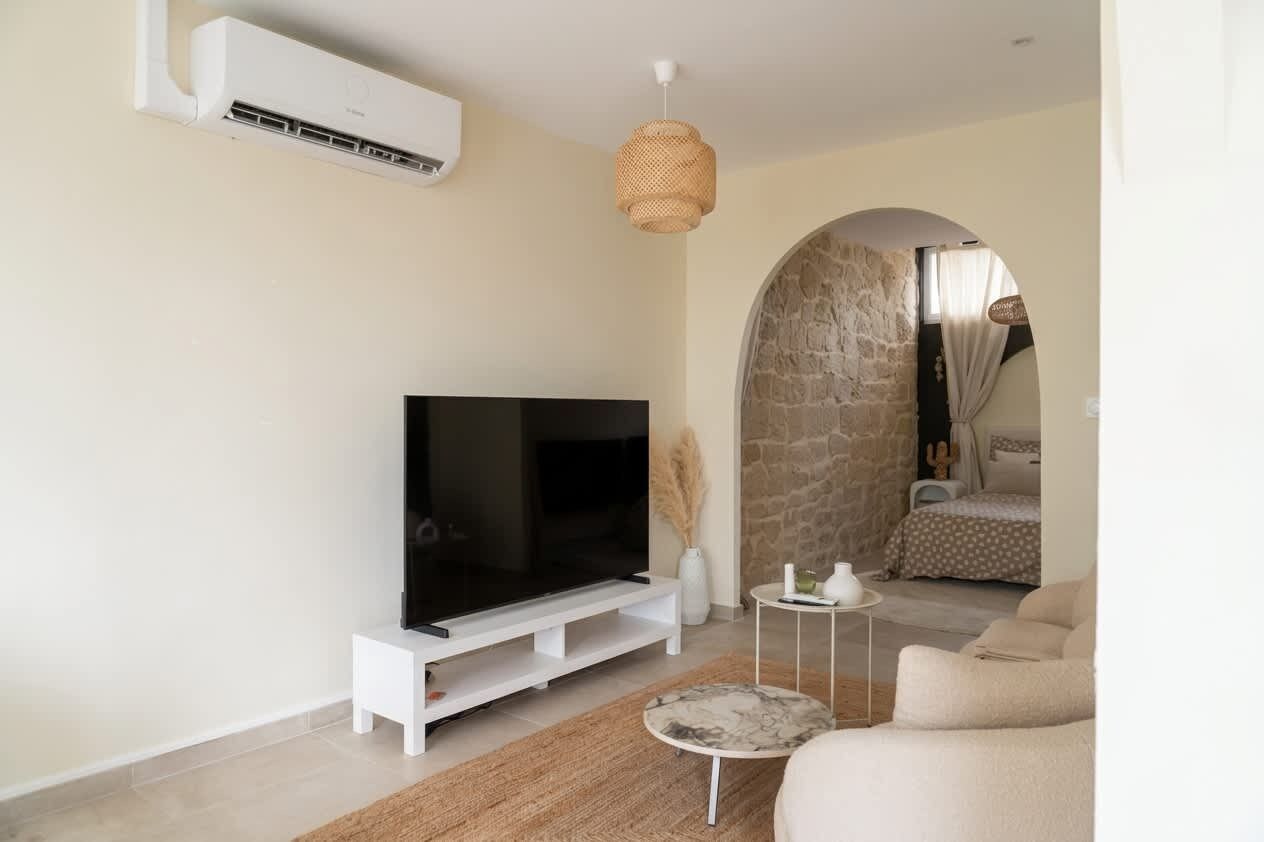Un salon moderne avec un téléviseur à écran plat posé sur un meuble blanc, un mur en pierre, un luminaire en fibres tressées et un lit recouvert d’une couvre-lit à pois.
