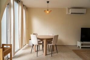 A dining area with a round table, chairs, a hanging light fixture, and a television on a stand. - Kalimera - Jacuzzi & Sauna (Oullins-Pierre-Bénite)
