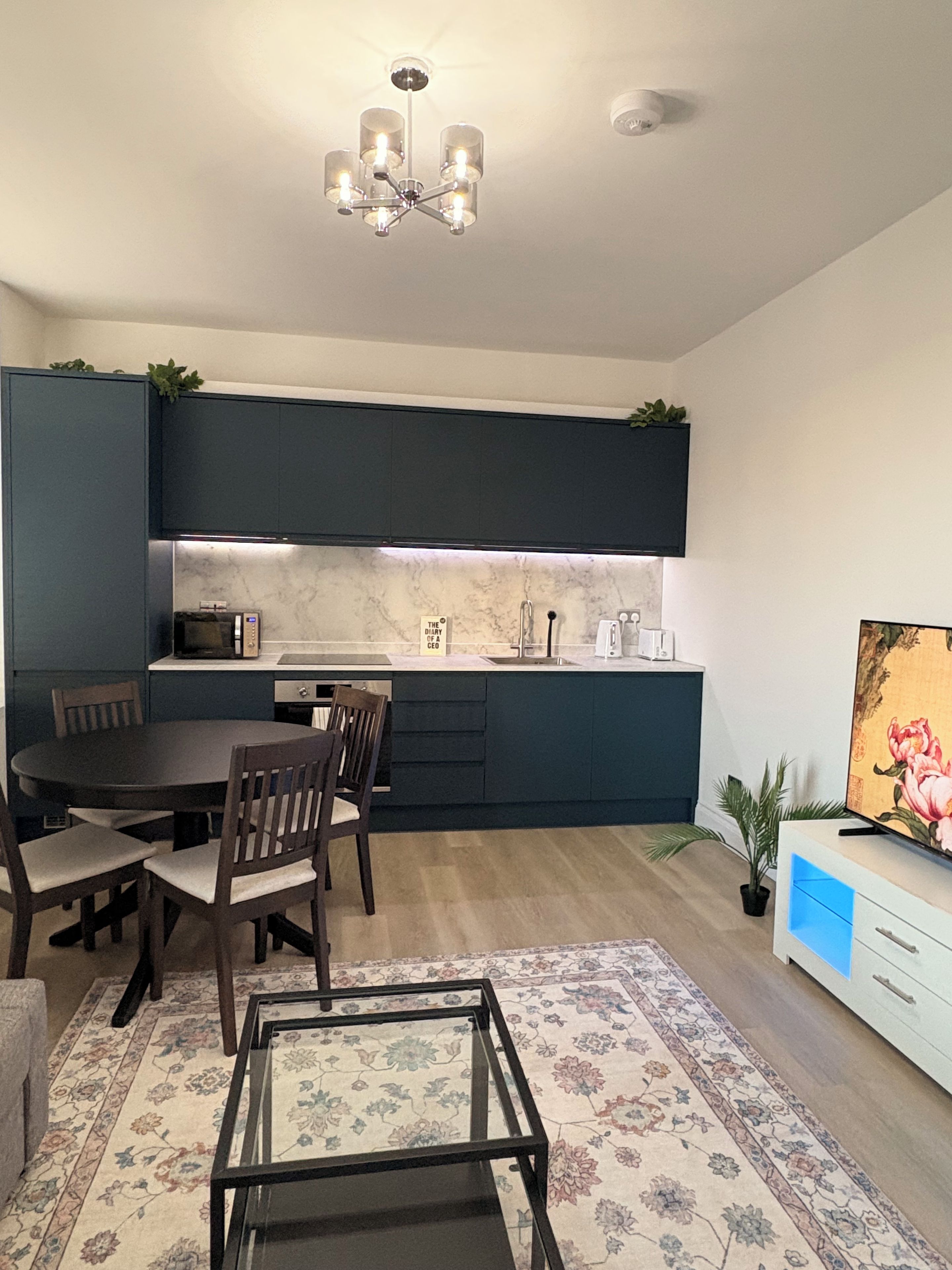 Een moderne keuken met donkere kasten, een eettafel met stoelen, een kroonluchter en een flatscreen televisie op een standaard.