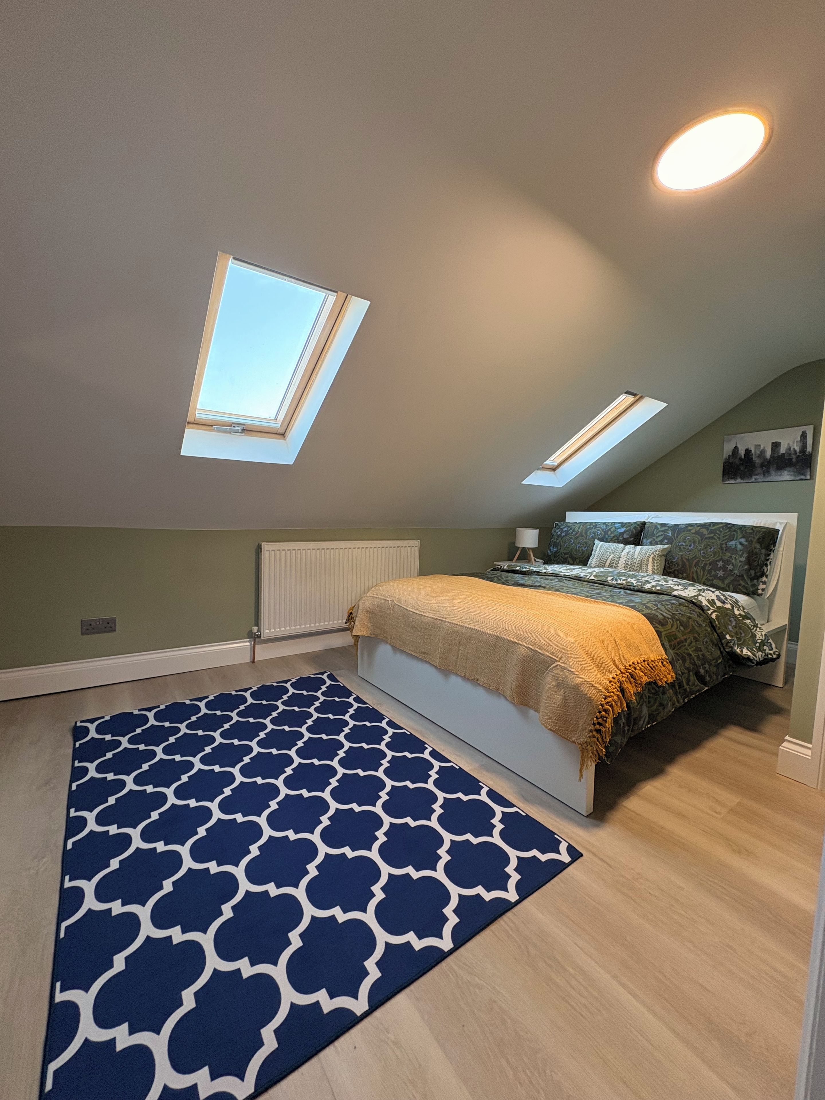 Une chambre avec un puits de lumière, un tapis à motifs, un lit avec une literie à motifs et un tableau représentant un paysage urbain accroché au mur.