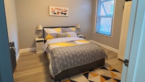 Ein Schlafzimmer mit Bett, Nachttischen, einem Fenster mit Jalousien, einer Heizung und einem Teppich mit geometrischem Muster.