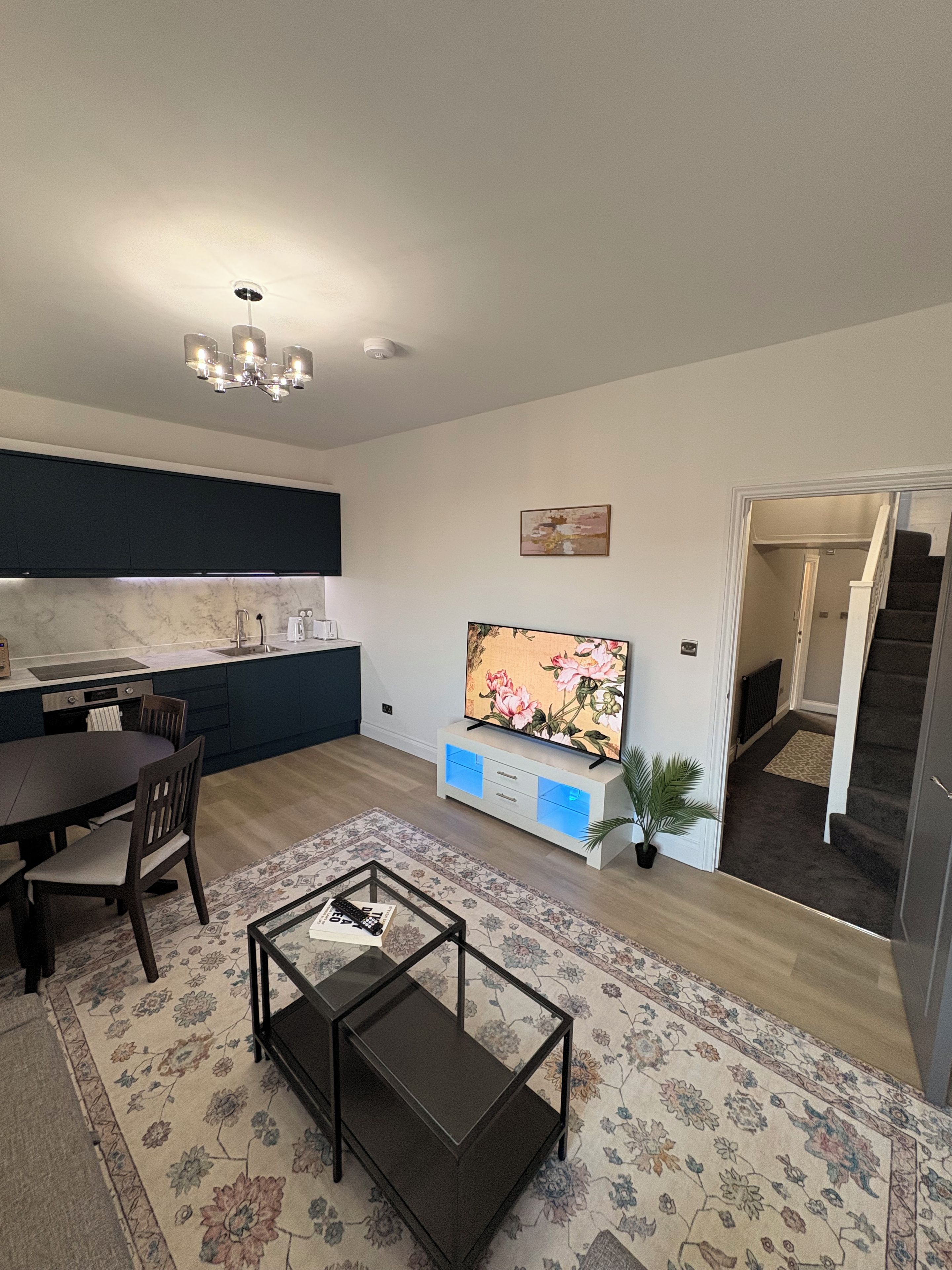 Une cuisine moderne avec des meubles de couleur sombre, un coin repas avec une table et des chaises, un téléviseur à écran plat posé sur un meuble blanc, et un escalier menant à l’étage supérieur.