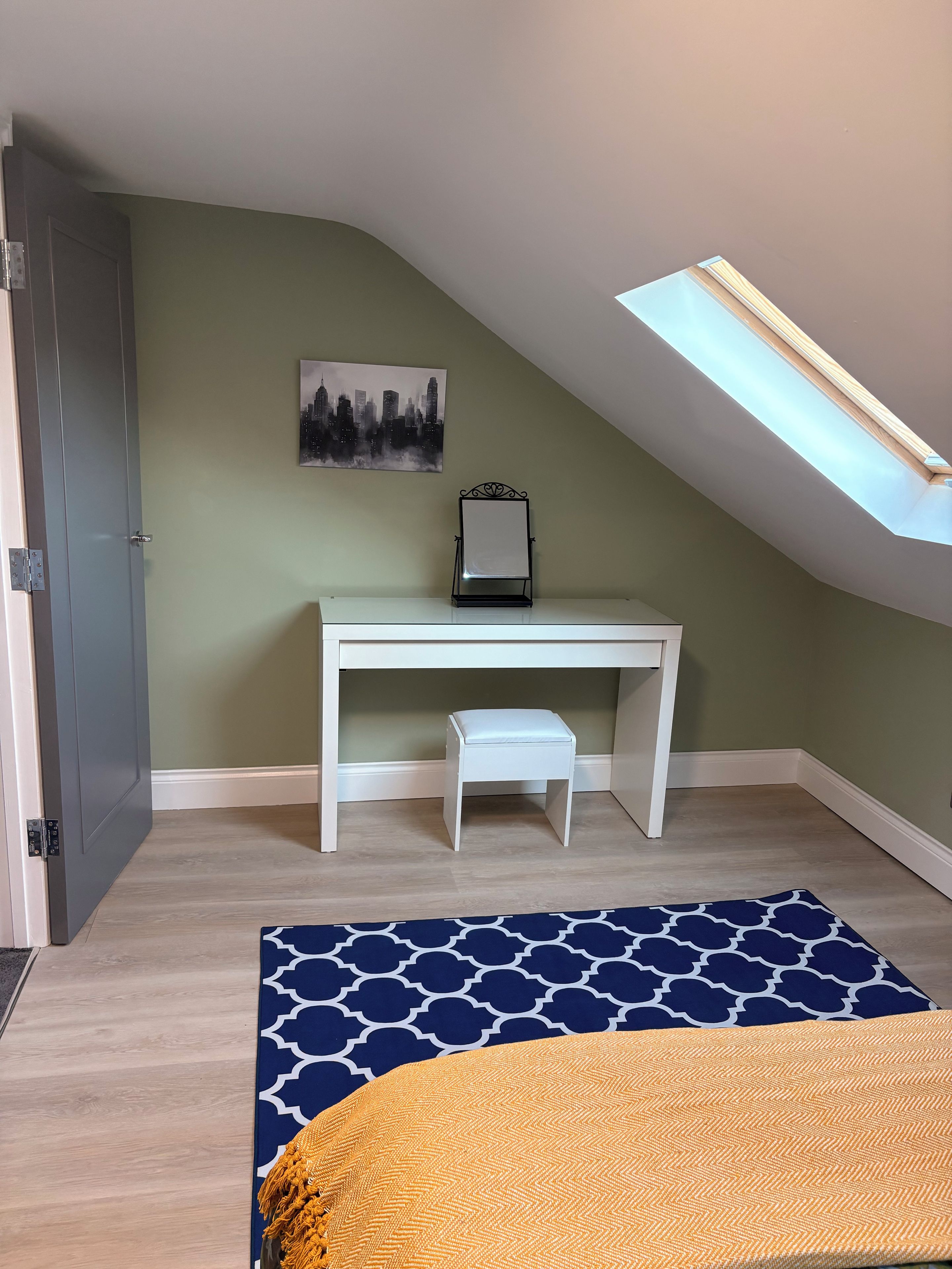 Une pièce avec un plafond en pente, un bureau avec une chaise, un petit tabouret, un tapis à motifs et un lit recouvert d’une couverture jaune.