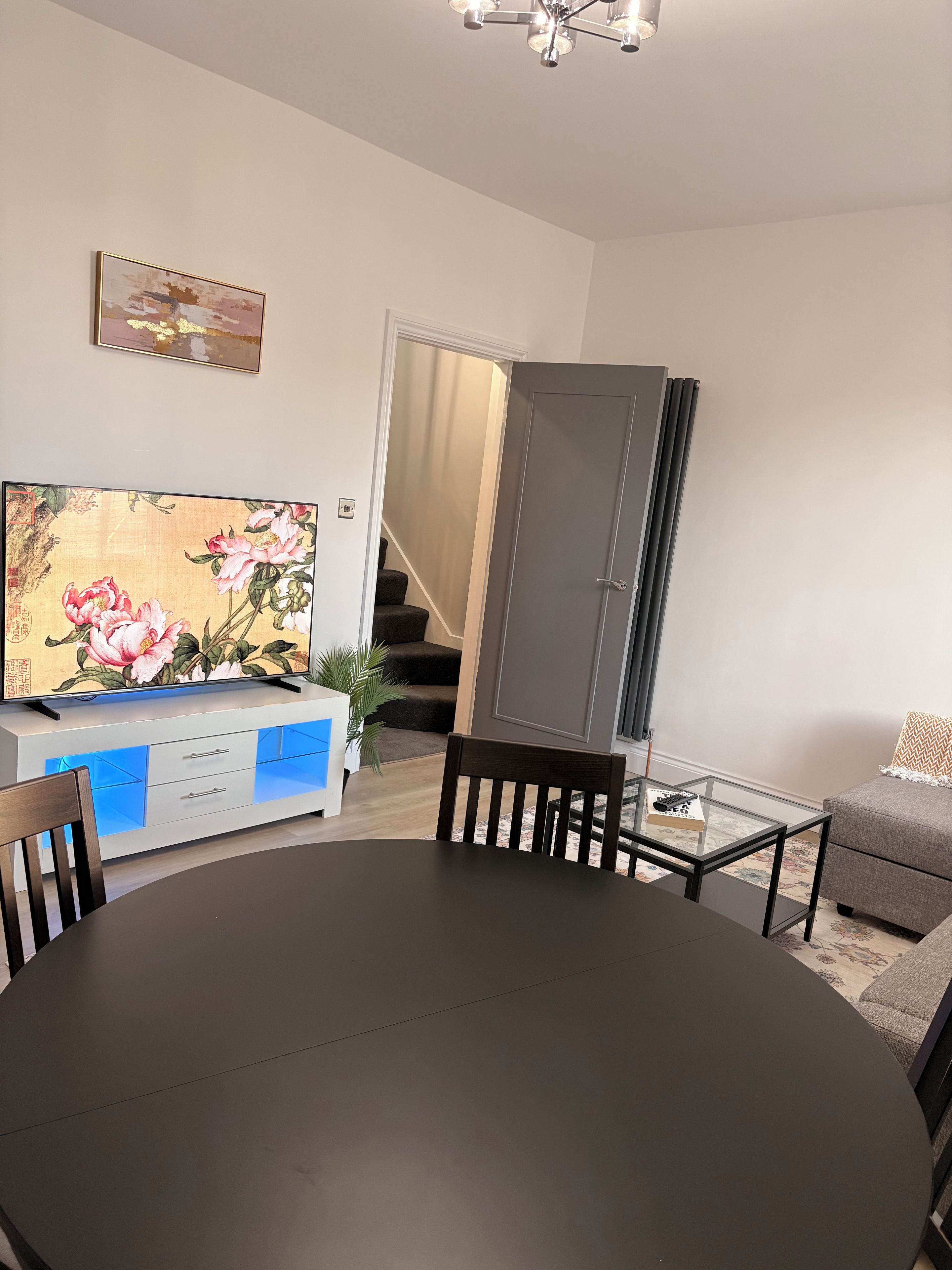 A dining area with a round table, chairs, a television on a cabinet, a sofa, and a staircase leading to another room.