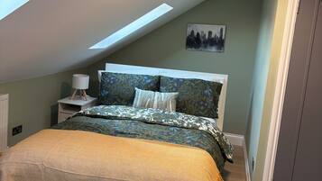 A bedroom with a skylight, a bed with patterned bedding, a nightstand with a lamp, and a cityscape painting on the wall.