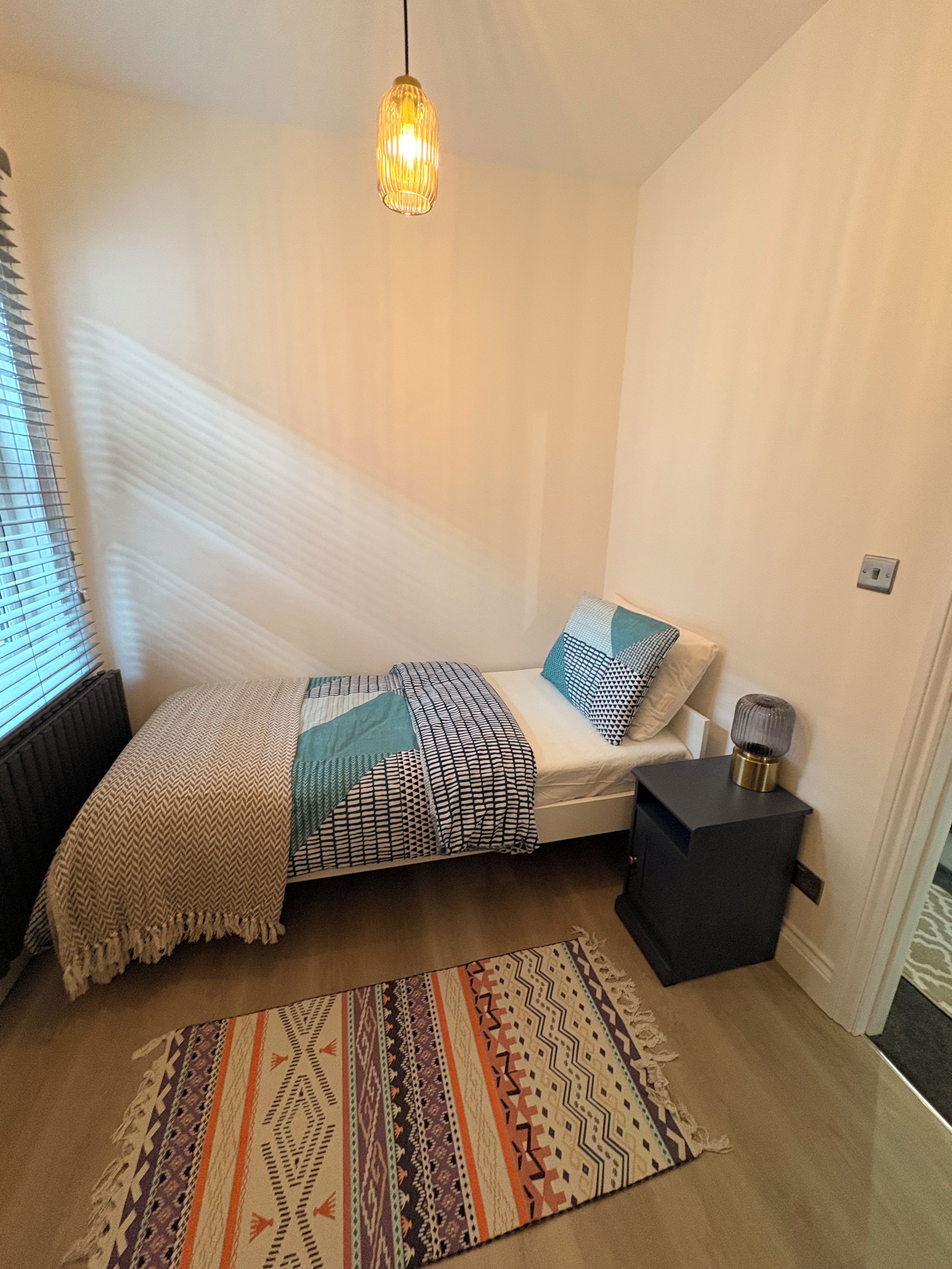 A small, well-lit bedroom with a bed, bedside lamp, and a patterned rug.