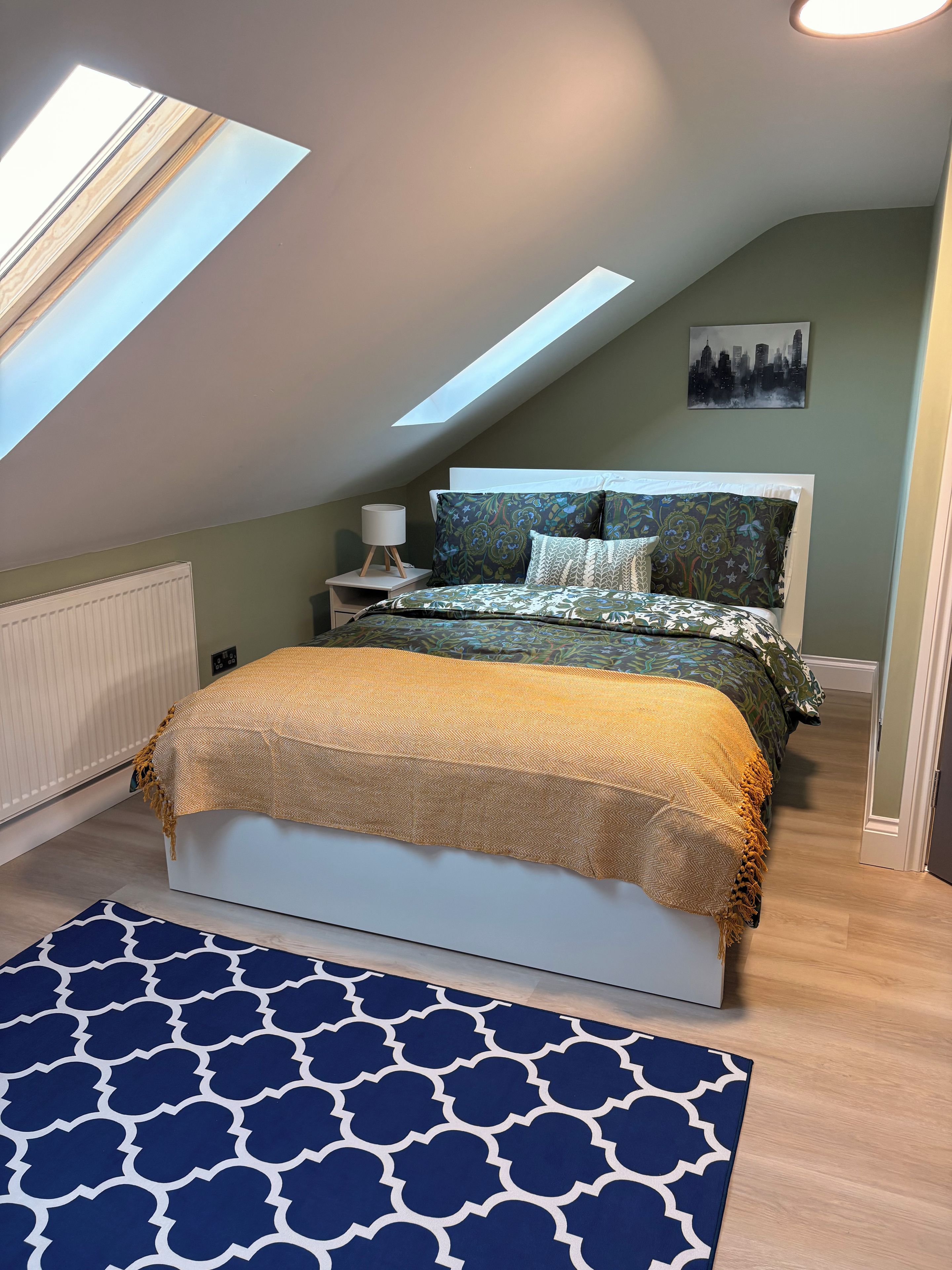 Een slaapkamer met dakraam, een bed met beddengoed met een patroon, een nachtkastje met een lamp en een stadsgezicht schilderij aan de muur.