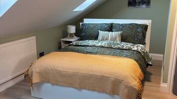Een slaapkamer met dakraam, een bed met beddengoed met een patroon, een nachtkastje met een lamp en een stadsgezicht schilderij aan de muur.