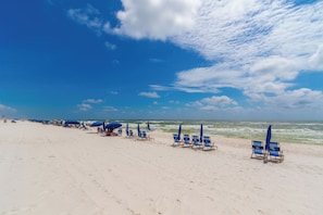 Ein Strand mit Reihen aus blauen Liegestühlen und Sonnenschirmen, einem sandigen Ufer und dem Meer im Hintergrund.