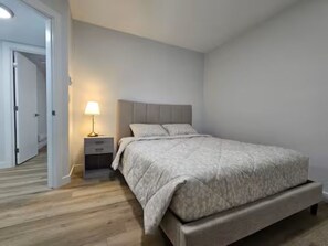 A bedroom with a bed, bedside table, lamp, and a door leading to another room. - Marise Tranquility - New Cozy 2-Bedroom Suite (Saskatoon)