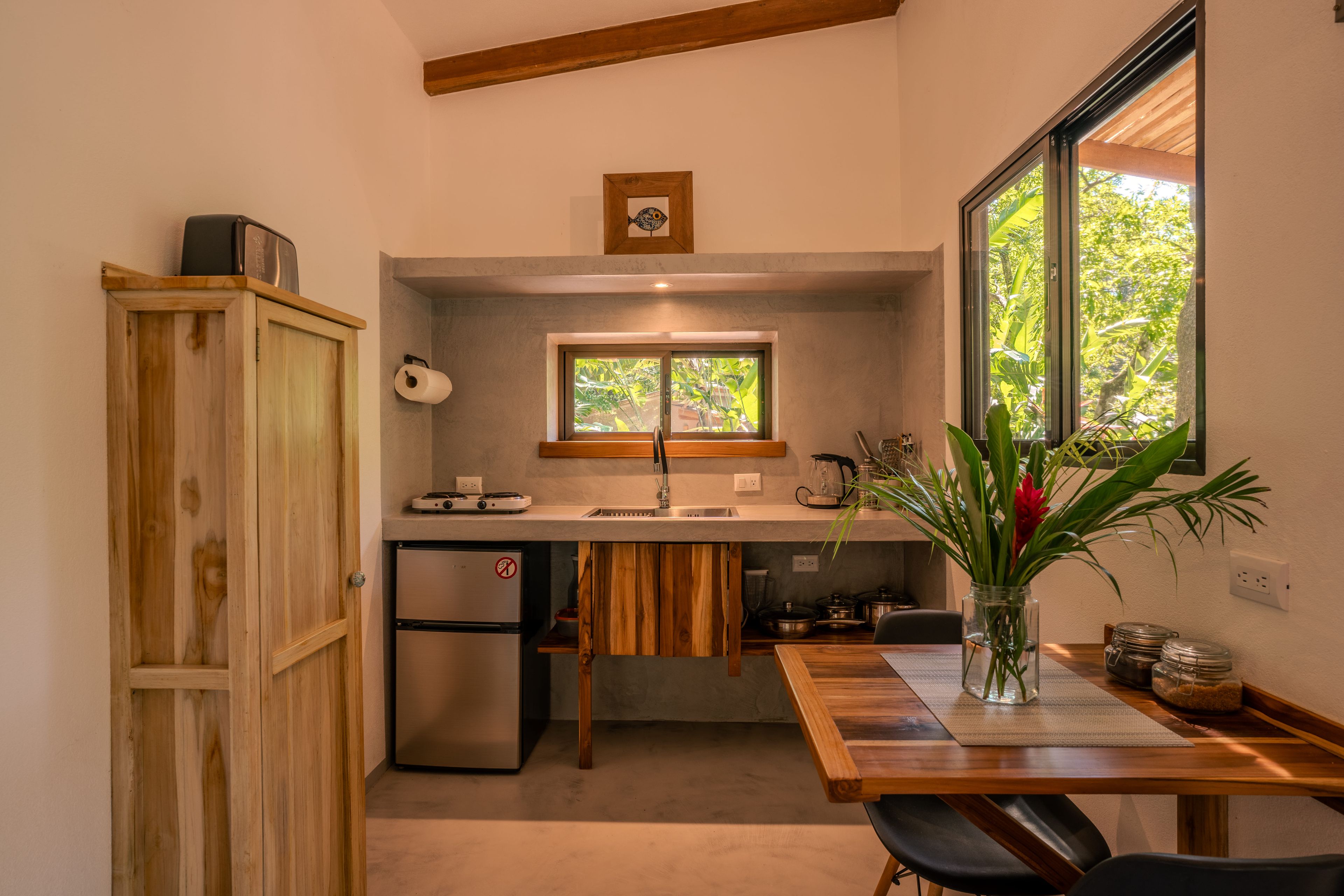 Une cuisine compacte avec un meuble en bois, un réfrigérateur encastré, une fenêtre donnant sur un espace verdoyant et un coin repas avec une table et des chaises.