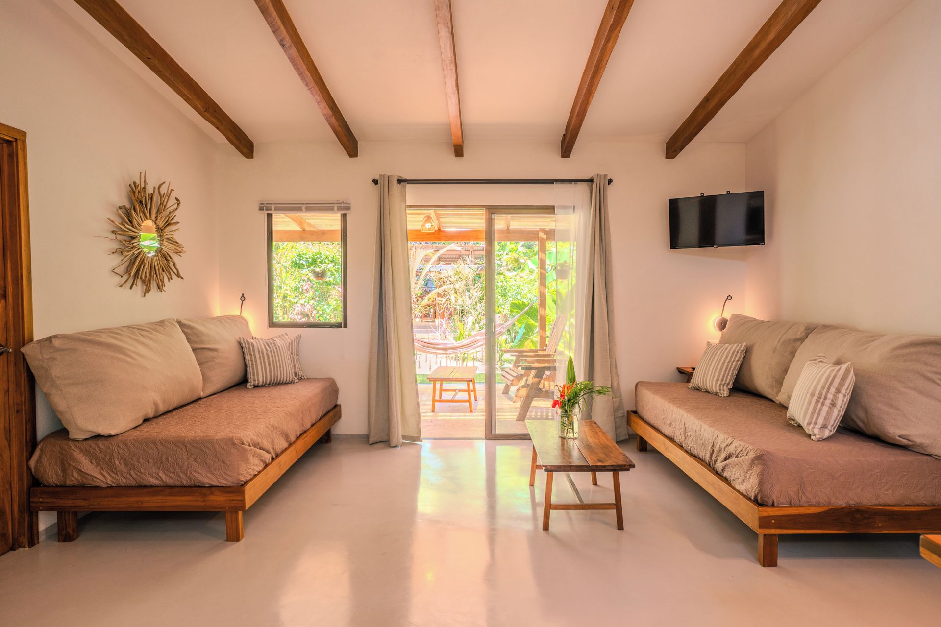 A room with two wooden sofas, a small table, a TV mounted on the wall, and a large sliding glass door leading to an outdoor area with greenery.