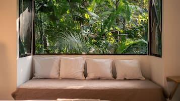 Una cama con colchón color beige y almohadas blancas, frente a una ventana con vistas a un follaje verde y tupido.