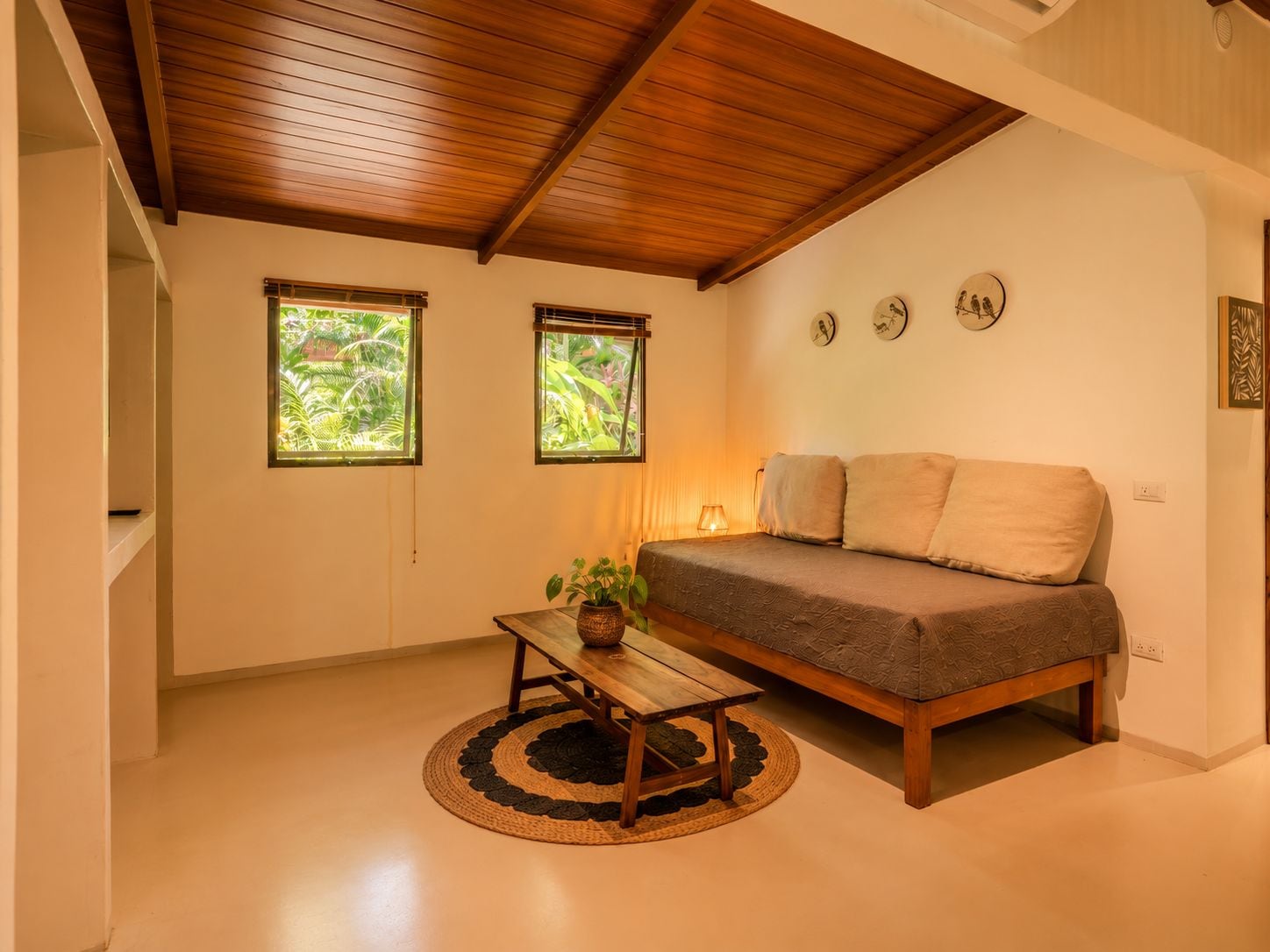 Una habitación acogedora con techo de madera, una cama, una mesita con una planta y dos ventanas con vistas a la vegetación.
