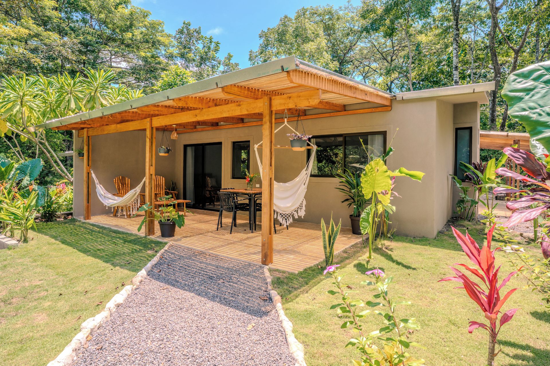 A covered outdoor patio with a table and chairs, surrounded by potted plants and a gravel path.