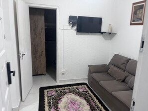 Uma sala de estar com sofá, televisão na parede e porta que dá para outro cômodo.