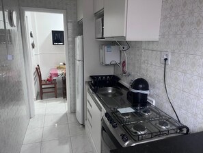 Cozinha com fogão a gás, geladeira e micro-ondas. Uma cadeira e uma mesa com toalha xadrez ao fundo.