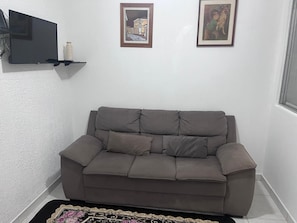 A grey sofa in a corner of a room with a white textured wall, a flat-screen TV mounted on the wall, and two framed pictures above the sofa. - Suitable for weekends and holidays, very close to the beach - Vila Guilhermina (Praia Grande)