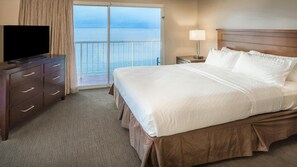 Habitación de hotel con cama, televisor, mesita de noche, lámpara y vista al mar desde el balcón.