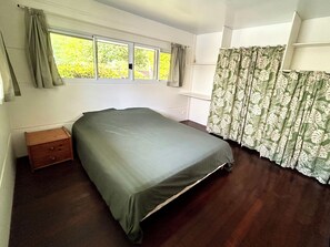A single bed with a grey bedspread in a room with a window, a small wooden nightstand, and a patterned curtain.