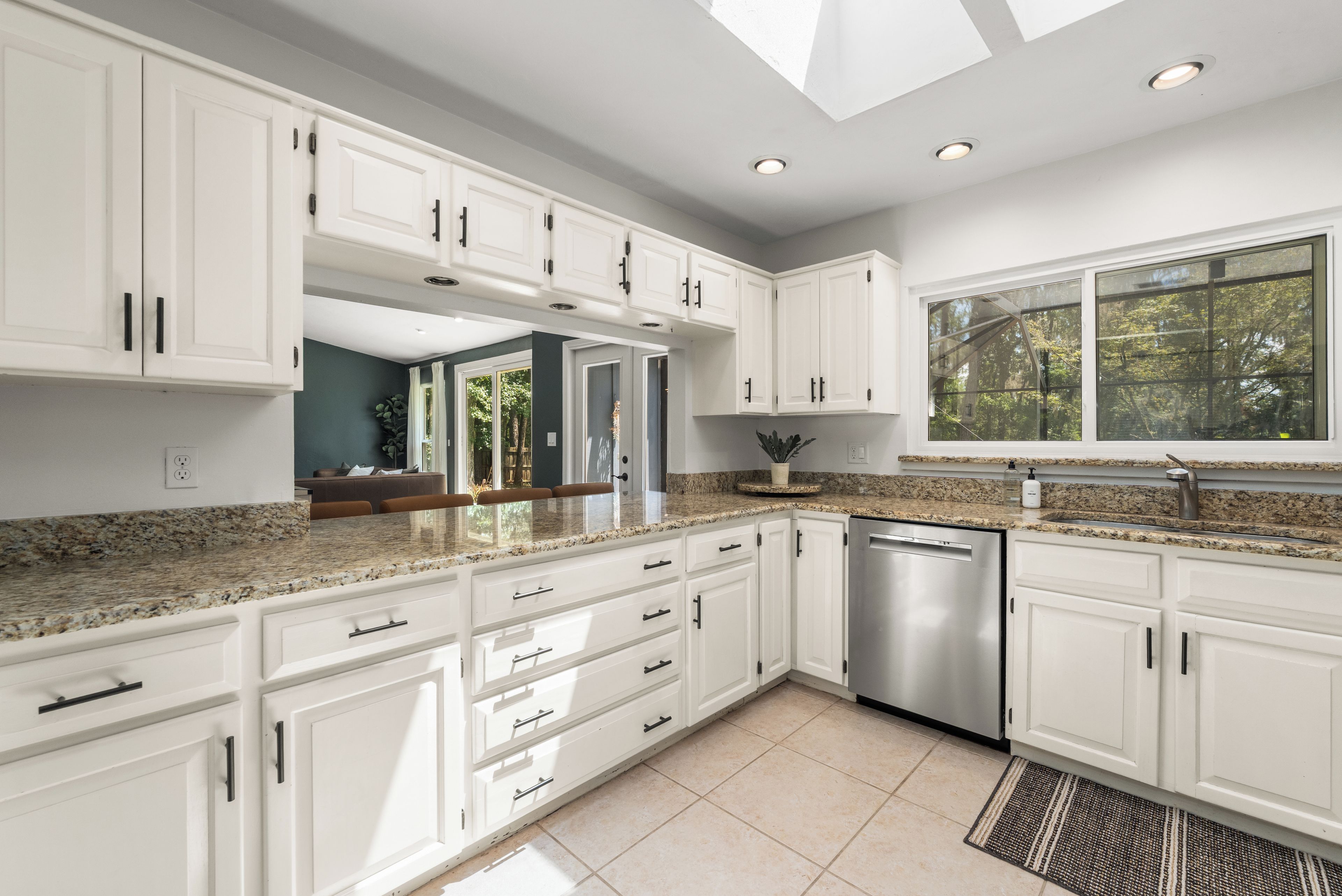 A modern kitchen with white cabinetry, granite countertops, a stainless steel dishwasher, and a large window providing natural light.
