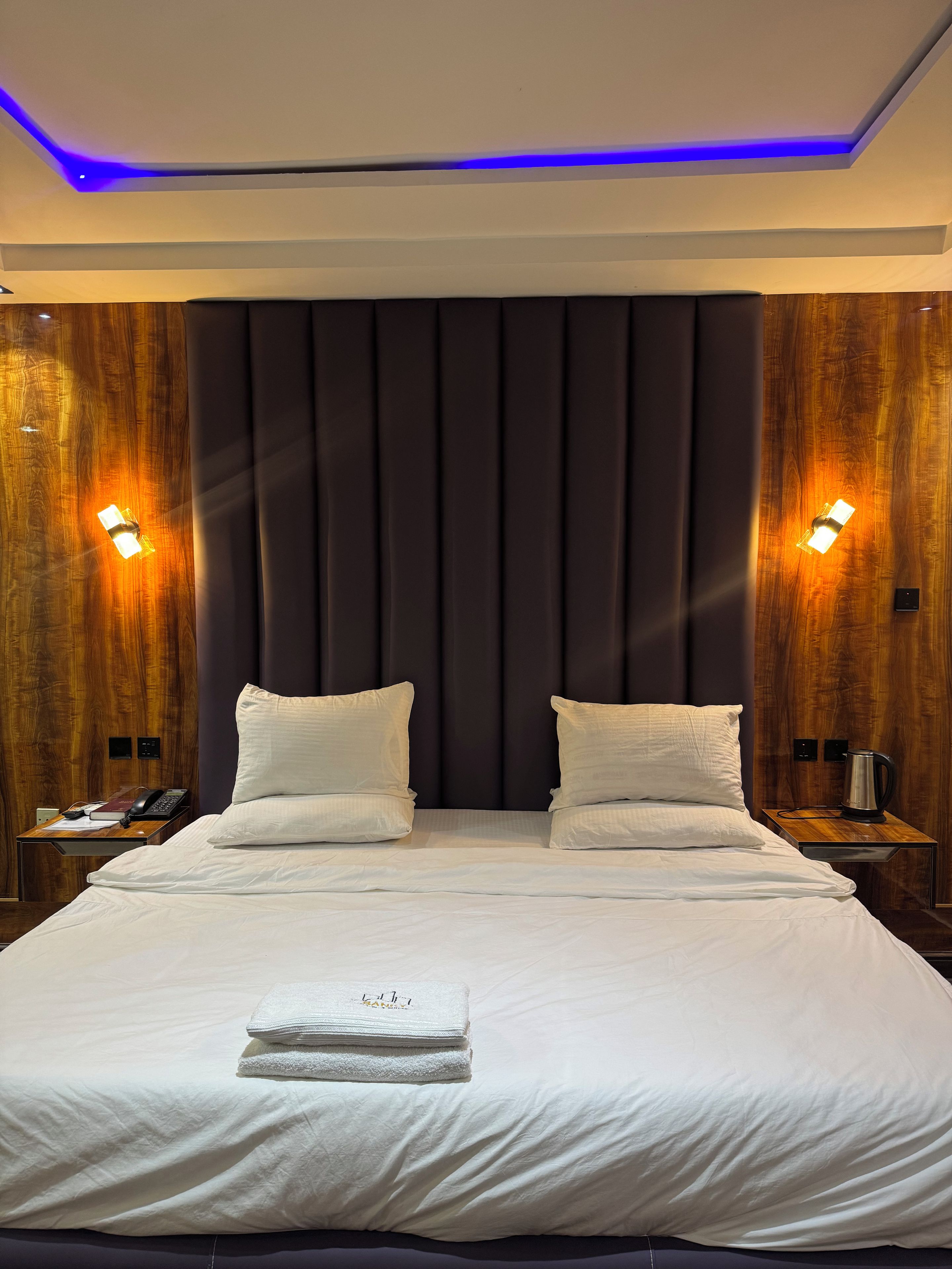 Quarto de hotel com uma cama grande, duas mesas de cabeceira, painel de cama de madeira e iluminação embutida na parede.