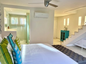 Une chambre à coucher comprenant un lit, un ventilateur de plafond, un appareil de climatisation, un escalier et une salle de bain avec une douche et un miroir.