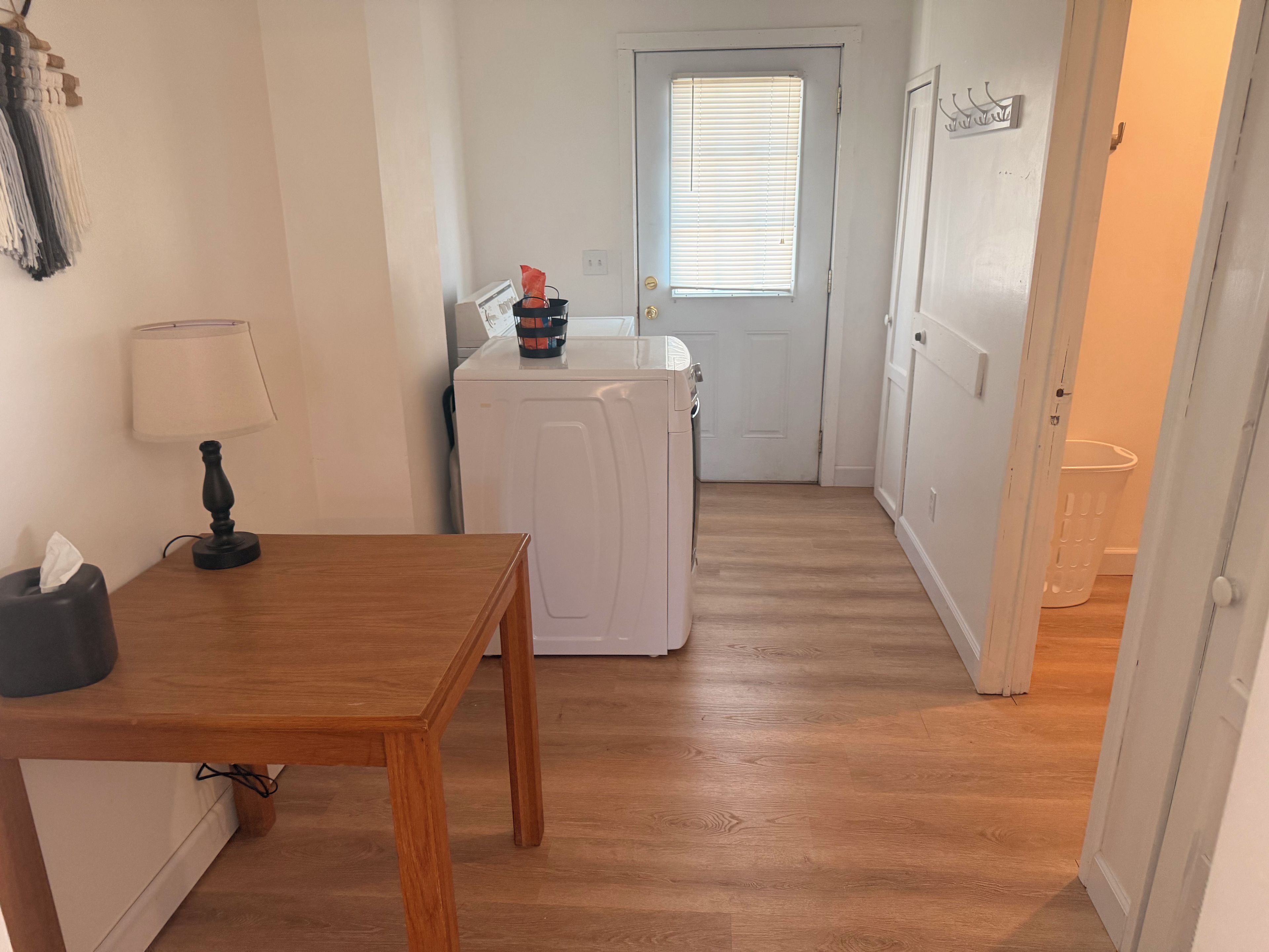 A compact kitchen with a washing machine, a table with a lamp, and a door leading to another room.