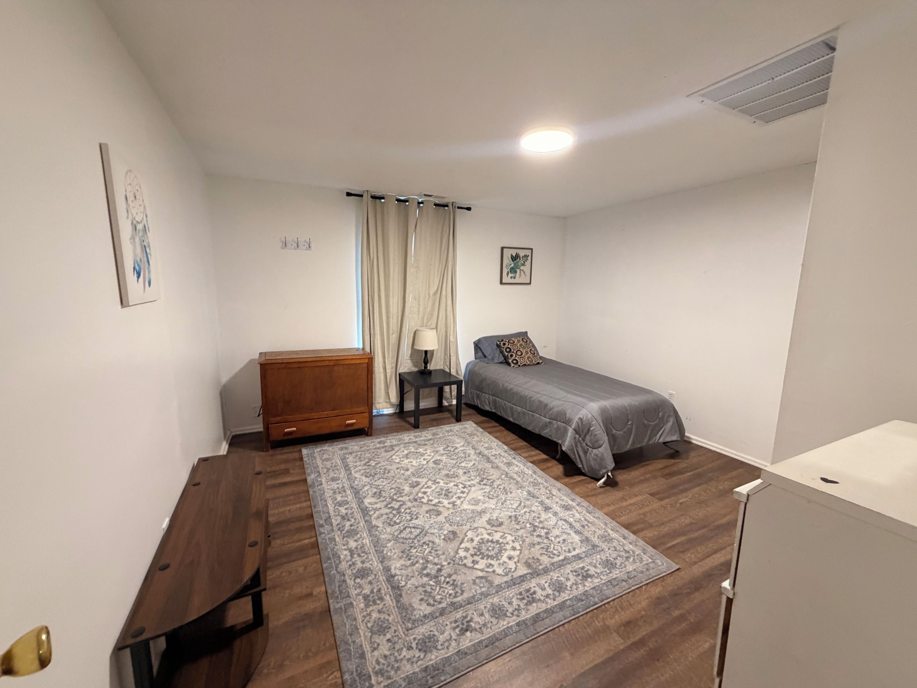 Allotjament petit i d'una sola habitació amb un llit, un taulell de fusta, una taula petita, una làmpada, una catifes i un armari blanc.