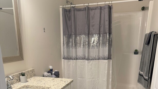 Een badkamer met een wit toilet, een wastafel met een granieten aanrecht, een douche met een grijs douchegordijn en een kleine potplant op het aanrecht.