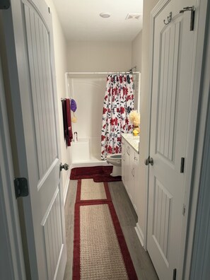 Un couloir étroit avec un espace douche, une porte dotée d’un porte-serviettes et une petite table avec une lampe.