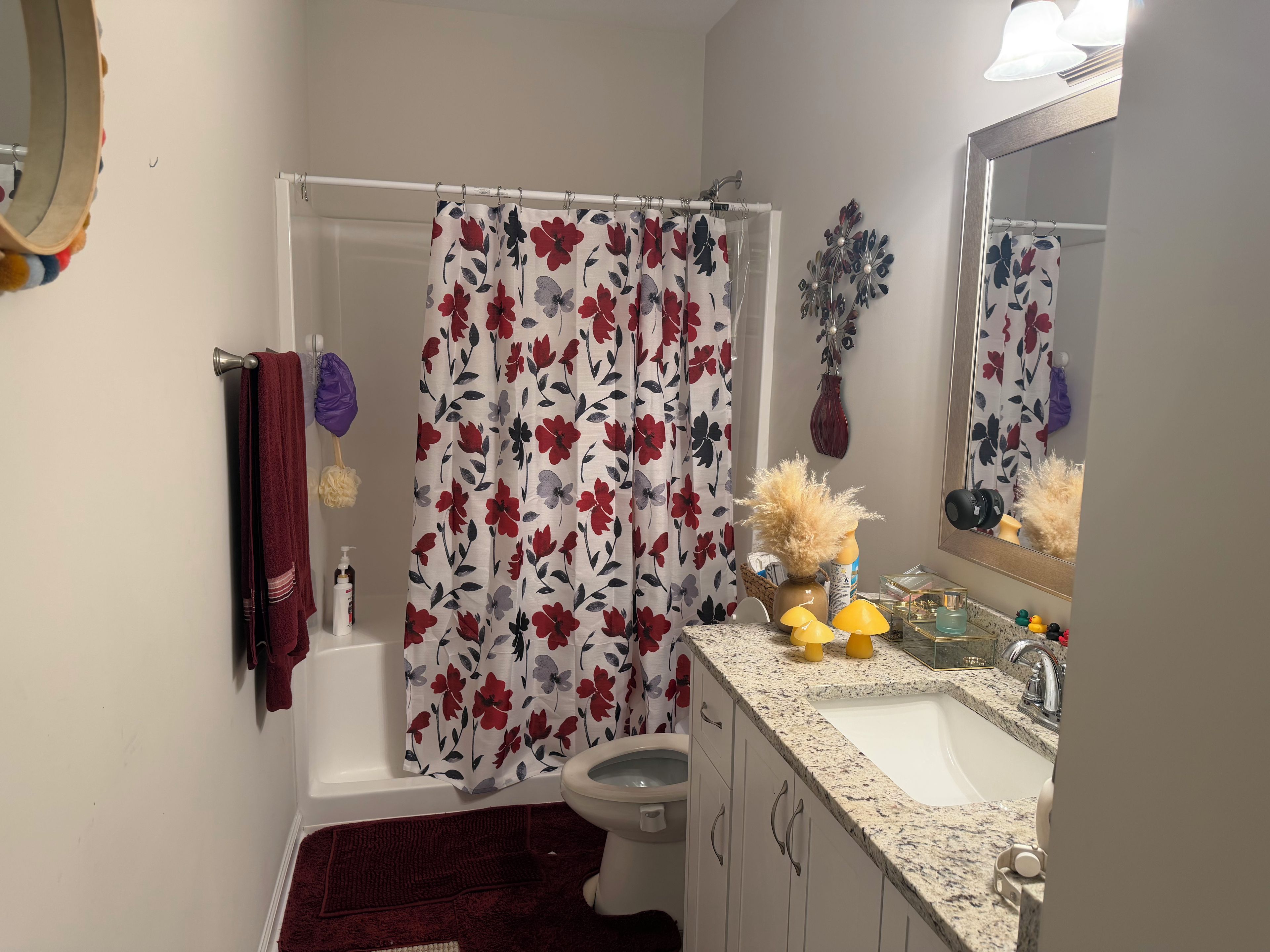 Un baño con bañera blanca, inodoro, lavamanos con encimera de granito y cortina de ducha estampada con motivos florales.