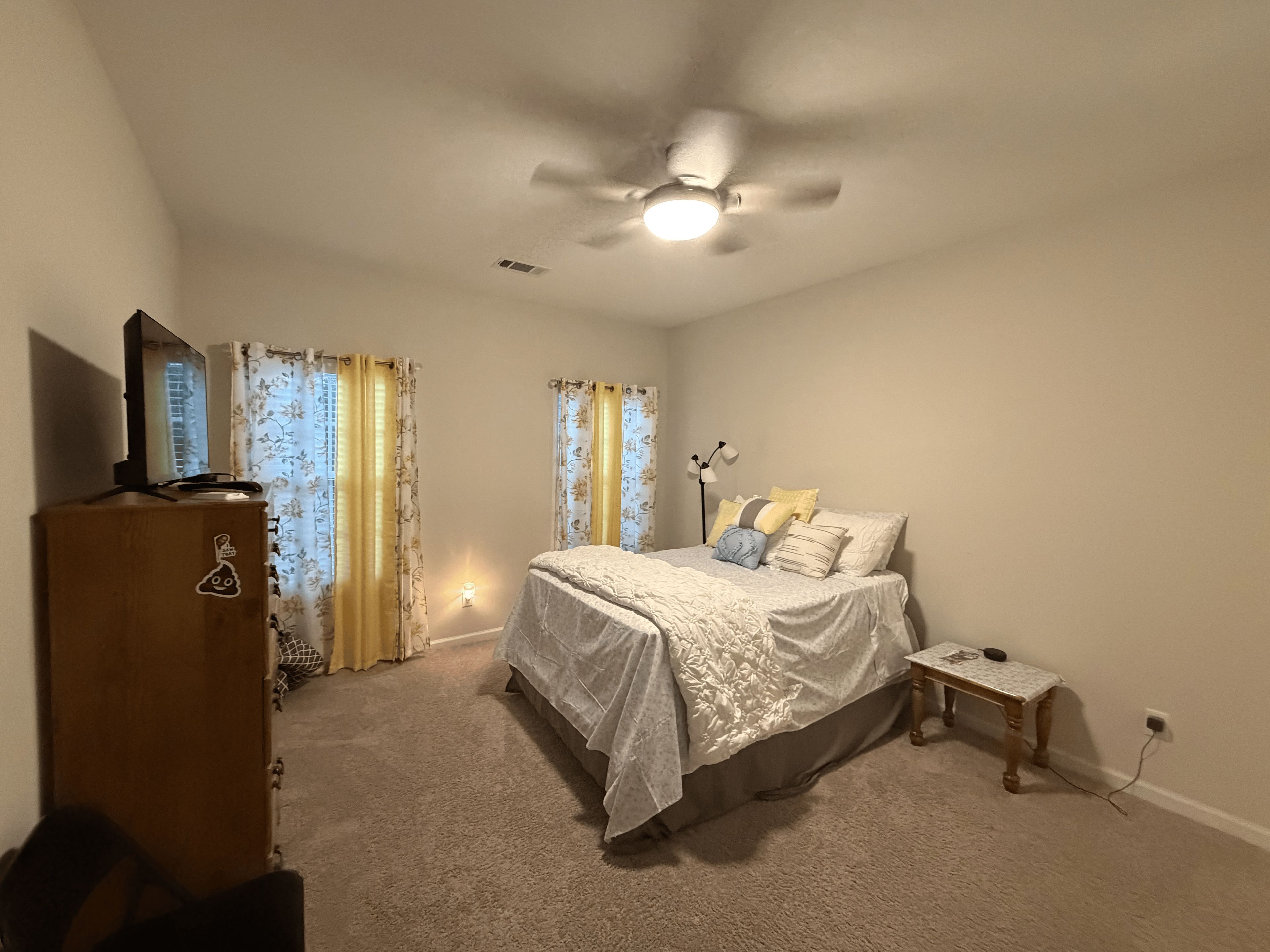 A bedroom with a bed, a nightstand, a desk, a ceiling fan, and a dresser.