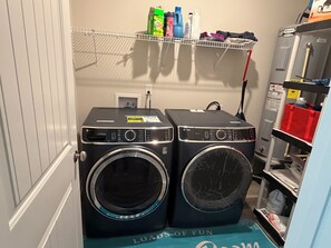 Two front-loading washing machines in a laundry room with a shelf above containing various cleaning supplies.