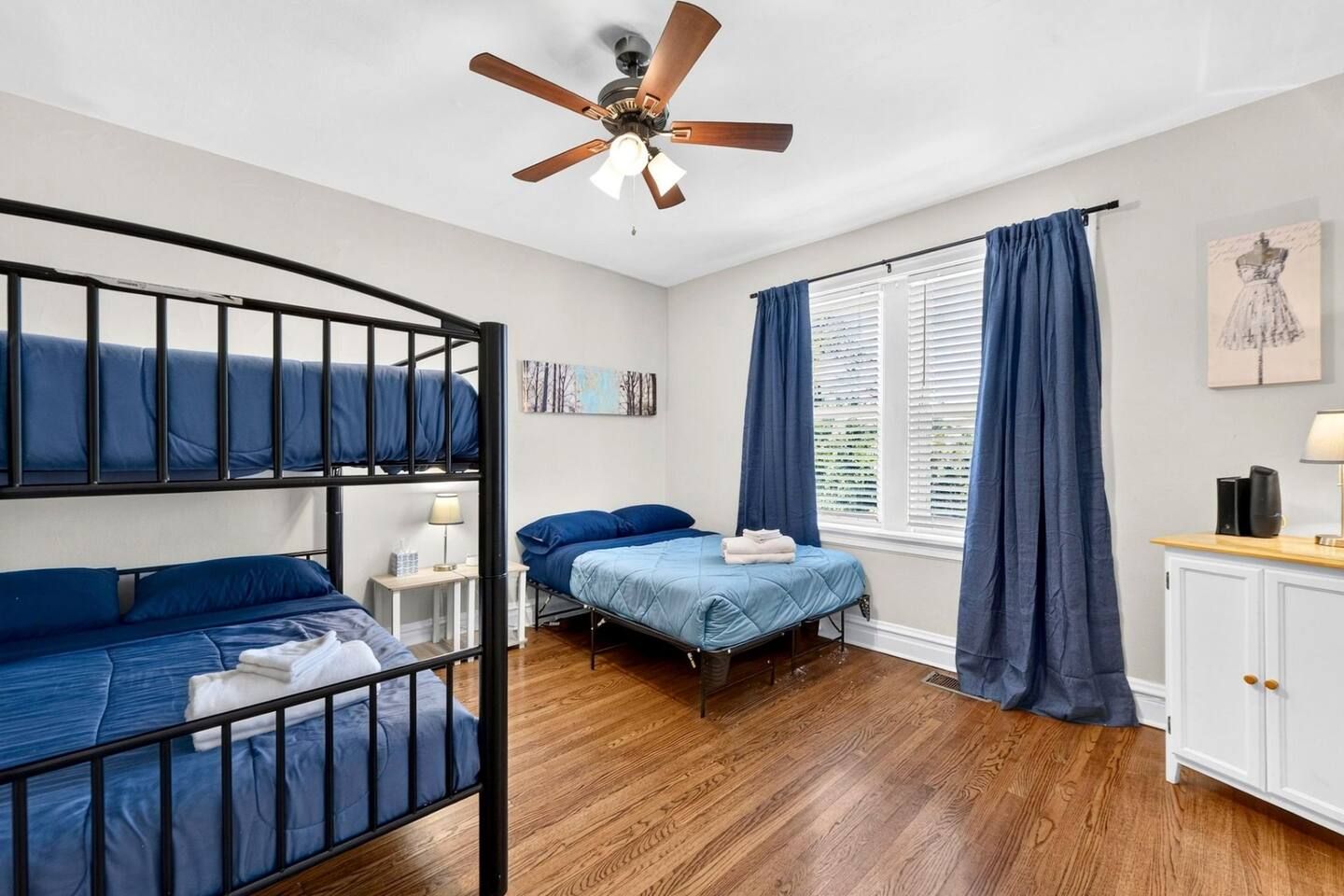 A bedroom with bunk beds, a single bed, a desk, a chair, a ceiling fan, and a window with curtains.