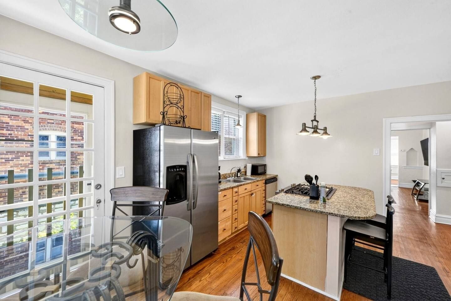 Una cocina moderna con refrigerador de acero inoxidable, armarios de madera, mesa de comedor de vidrio y vista a un edificio de ladrillo.