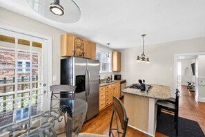 A modern kitchen with a stainless steel refrigerator, wooden cabinets, a glass dining table, and a view of a brick building outside.