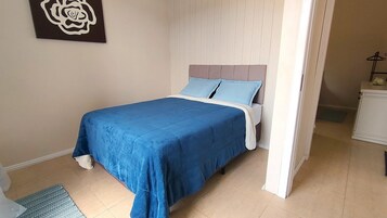 Una cama con colcha y almohadas azules, cabecera blanca y una mesita de noche con lámpara.
